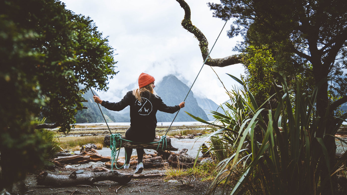 A woman from behind dressed in a black Saltwater Sketch Print Crew swinging on a swing in Milford Sound New Zealand with the famous Mitre Peak in the background.