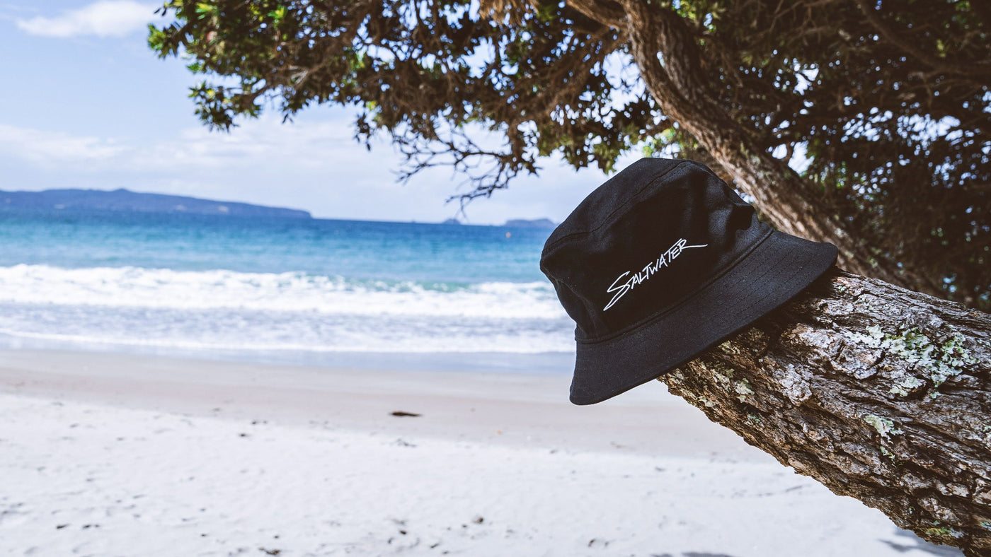 Saltwater bucket hat in navy is hanging on a tree. In the background you see one of the beautiful beaches of the coromandel.