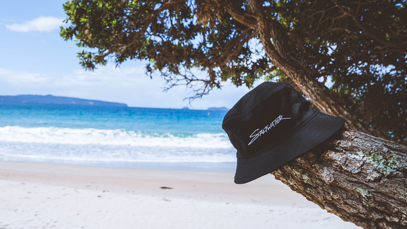 A navy Saltwater Bucket Hat laying on a branch of a tree with the beach in the background.