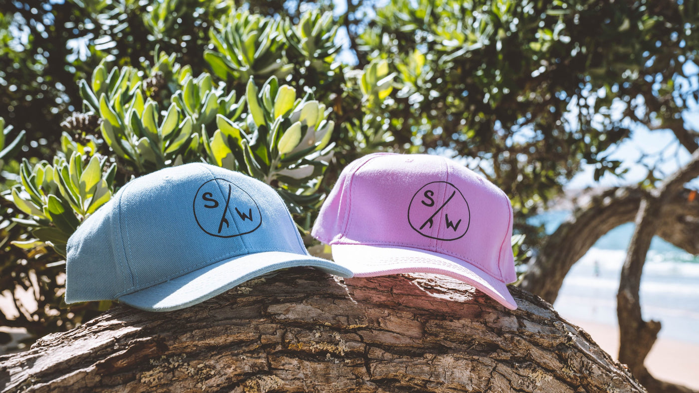 Two Saltwater Kids Caps in blue and pink laying on a branch of a tree with the beach in the background.