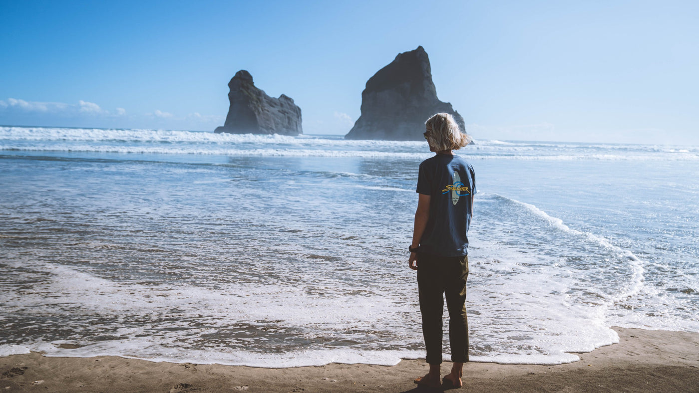 A man from behind dressed in a petrol blue Saltwater Board Print Shortsleeve Tee standing on a New Zealand Beach facing the waves rolling in.