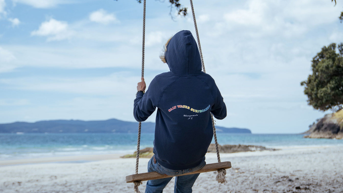 A man from behind dressed in a petrol blue Saltwater Original Hooded Sweatshirt swinging on a swing at Otama Beach in New Zealand.