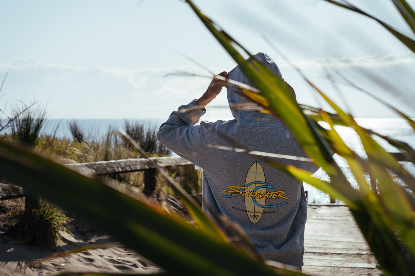 A woman from behind putting the hood of her Saltwater Board Print Hooded Sweatshirt on. She is standing on a walkway to the beach which is visible in the background.