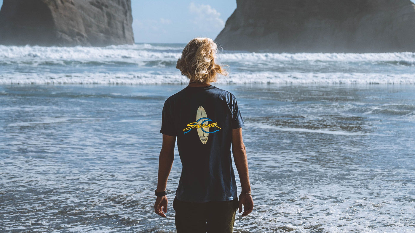 A man from behind dressed in a petrol blue Saltwater Board Print Shortsleeve Tee walking towards the waves on a New Zealand Beach.