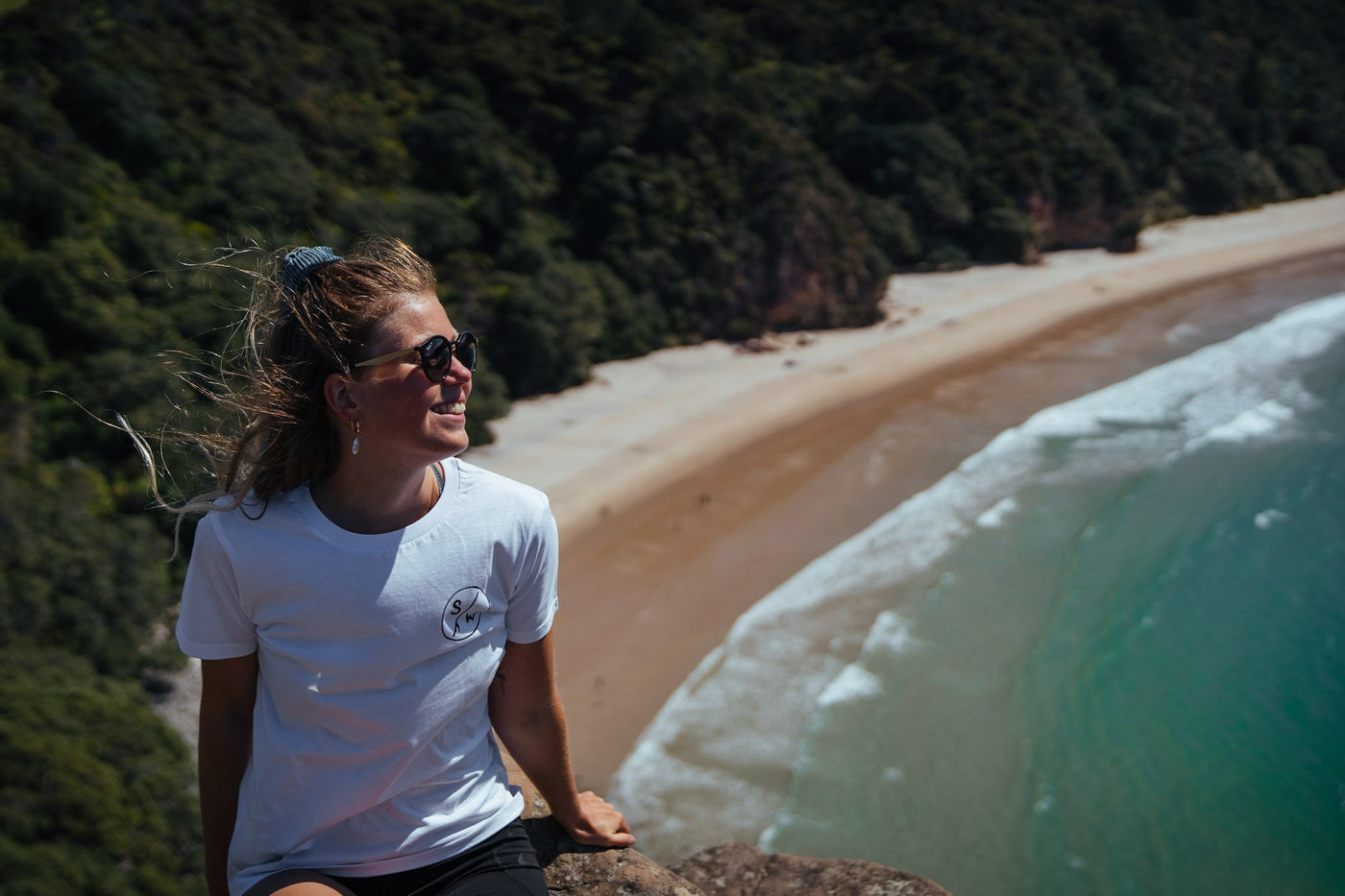 A smiling woman in a white Saltwater Sketch Print short sleeve tee sits on a rock at the viewpoint for New Chums beach which is visible in the background.