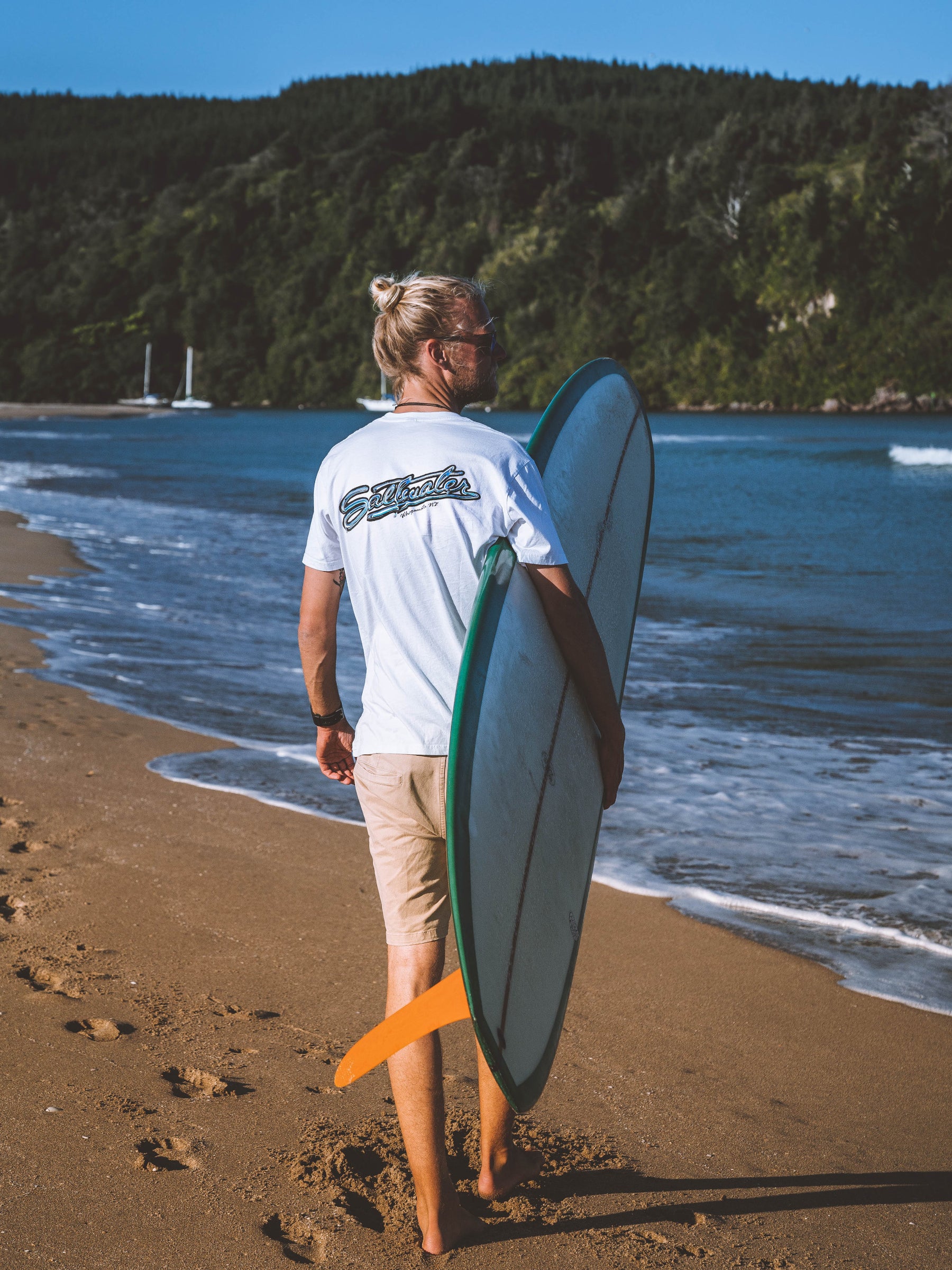 A men walking with his saltwater surfboard on the beach in Whangamata and wears his saltwater tubular tee.